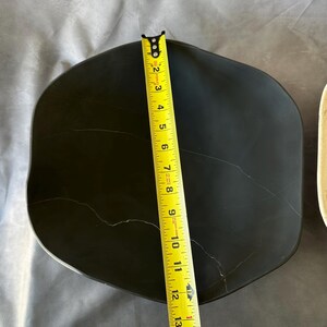 May include: A black, irregularly shaped plate with a yellow measuring tape showing a length of approximately 12 inches. The plate has visible cracks and a slightly reflective surface.