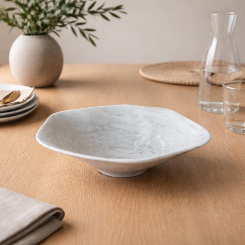 May include: A white marble-patterned serving bowl with a unique, geometric shape. The bowl is set on a light wood table, alongside a vase with greenery, a water carafe, and glassware. The bowl's design adds a touch of elegance to any dining setting.