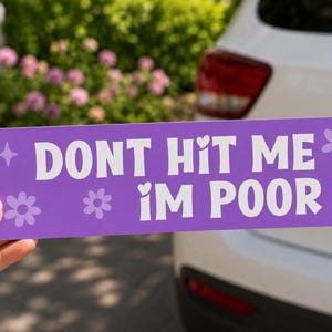 May include: A purple bumper sticker with white text that reads "DON'T HIT ME I'M POOR." The sticker has flower and star graphics. The sticker is being held in front of a white car.
