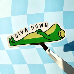 May include: A sticker featuring a cartoon frog in a relaxed pose, with the words "DIVA DOWN" above it. The frog is green and yellow with black outlines. The sticker is held by a pair of tweezers against a blue and white checkered background.
