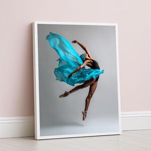 May include: A framed print showing a ballet dancer mid-leap. The dancer is dressed in a black leotard and a flowing turquoise fabric. The image is set against a grey backdrop, with the frame leaning against a pink wall, creating a sense of movement.