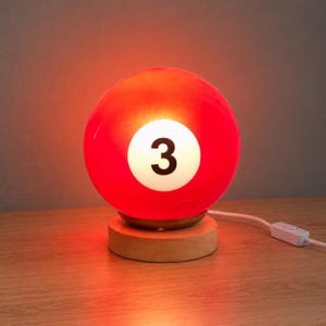 May include: A spherical red lamp shaped like a billiard ball with a white circular center featuring the black number 3. The lamp is mounted on a circular light-colored wooden base and includes a white power cord with a small inline switch.