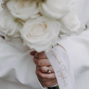 Könnte beinhalten: Ein Strauß weißer Rosen, umwickelt mit einem weißen Band mit goldener Schreibschrift. Das Band zeigt Namen in fließender Schrift. Am Finger sind zwei goldene Ringe sichtbar, einer davon ein Solitär-Diamant-Verlobungsring.