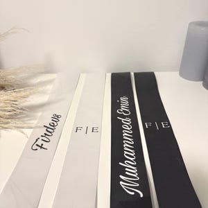 May include: White and black satin ribbons with custom text. One ribbon reads "Firdevs" in a script font. Another reads "Mohammed Emin" in a script font. Both have "F|E" printed on them. Ideal for personalized gifts or event decorations.