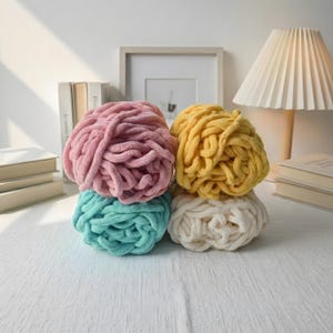 May include: Four balls of chunky yarn in pastel shades: pink, yellow, teal, and cream. The yarn is stacked on a white surface, with a lamp, books, and a framed picture in the background. The yarn appears soft and plush.