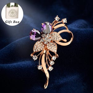 May include: A rose gold-toned metal brooch shaped like a flower, featuring two large, oval-cut purple crystals, smaller clear pavé-set rhinestones on the petals, and delicate metal stems. Includes a white gift box with the text Gift Box.
