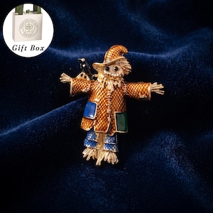 May include: A gold-tone metal brooch shaped like a scarecrow wearing a textured orange jacket with blue and green patches, blue patterned trousers, and a matching hat. A small black bird sits on the scarecrow's shoulder. Includes a gift box.