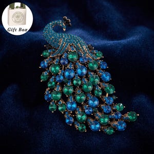 May include: A peacock-shaped brooch featuring a metallic gold-tone base encrusted with numerous round blue and green rhinestones. The design includes a curved neck and a fanned tail composed of layered, circular crystals in varying shades of jewel tones.