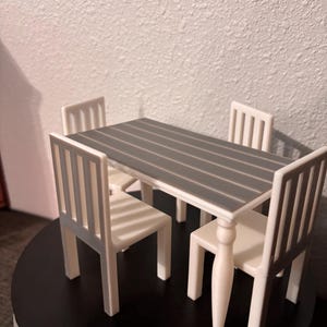 Puede incluir: Un juego de comedor en miniatura con una mesa rectangular con una tapa gris a rayas y cuatro sillas blancas. La mesa tiene cuatro patas blancas. Las sillas tienen un diseño de respaldo de listones. El juego se muestra sobre una superficie negra.