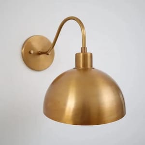 Könnte beinhalten: Messingfarbene Wandleuchte mit gebogenem Arm und kuppelförmigem Schirm. Die Leuchte ist an einer runden Grundplatte befestigt. Das Leuchtmittel hat ein Vintage- oder Industriedesign.