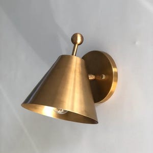 May include: A brass-colored wall sconce with a conical shade and a round backplate. The sconce has a small bulb inside the shade and a decorative ball on top. The fixture is mounted on a white wall.