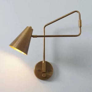 Könnte beinhalten: Eine messingfarbene Wandleuchte mit konischem Schirm und verstellbarem Arm. Die Lampe hat einen kreisförmigen Sockel und einen rechteckigen Arm, der sich vom Sockel aus erstreckt. Der Schirm ist nach unten abgewinkelt, und das Innere ist hellgoldfarben.
