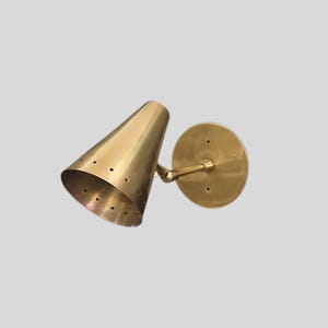 Könnte beinhalten: Messing-Wandleuchte mit konischem Schirm und kreisförmiger Rückplatte. Der Schirm hat kleine Perforationen in der Nähe der Öffnung. Die Leuchte ist aus Messing gefertigt und hat eine warme, metallische Oberfläche. Das Design ist minimalistisch und modern.