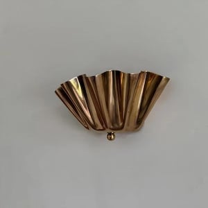 Könnte beinhalten: Eine goldfarbene Wandleuchte mit einem wellenförmigen, fächerartigen Design. Die Leuchte hat ein kleines, rundes Dekorationselement am unteren Ende. Die metallische Oberfläche reflektiert das Licht und erzeugt eine warme Atmosphäre. Dieses dekorative Stück ist ideal.