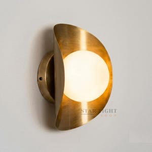 Könnte beinhalten: Eine Wandleuchte mit einem halbkreisförmigen Metallschirm aus gebürstetem Messing und einer zentralen, kugelförmigen weißen Glaskugel. Die Leuchte verfügt über einen runden Sockel und der Schriftzug STAR LIGHT SHOP ist unten rechts aufgedruckt.