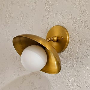 Op de afbeelding: Een messingkleurige wandlamp met een witte bolvormige lamp. De lamp heeft een gebogen, komvormige kap en een ronde achterplaat. De armatuur is gemonteerd op een getextureerde, gebroken witte muur. Ongeveer 20 cm.