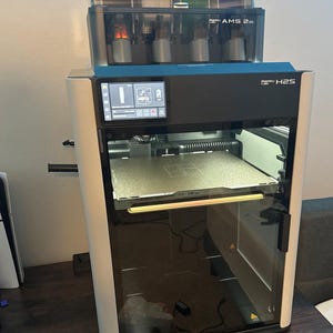 May include: A 3D printer with a black and white exterior, featuring a clear door and a digital display. Above it sits a multi-spool filament system with various coloured filaments. The printer is labelled "Bambu Lab H2S" and "AMS 2".
