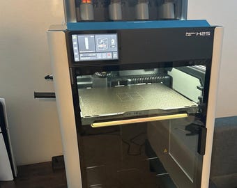 May include: A 3D printer with a black and white exterior, featuring a clear door and a digital display. Above it sits a multi-spool filament system with various coloured filaments. The printer is labelled "Bambu Lab H2S" and "AMS 2".