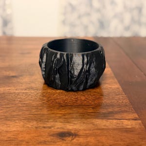 May include: A black, textured ring-shaped object with a rough, uneven surface. The object is made of a dark material and sits on a wooden surface. The ring has a hollow center and appears to be a decorative item.
