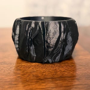 Op de afbeelding: Een zwarte, getextureerde, ronde container met een ruwe, organische uitstraling. De container staat op een houten oppervlak. De buitenkant van de container heeft een gekreukeld, ongelijkmatig oppervlak.