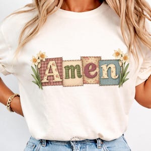 May include: Cream-colored t-shirt featuring the word "Amen" in a patchwork design. Each letter is on a different square with floral and patterned backgrounds. Daffodil illustrations flank the word. The shirt is a casual, everyday wear item.