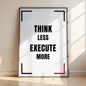 May include: A framed poster with the text "THINK LESS EXECUTE MORE" in bold black letters on a white background. The frame is black, with a red accent in the lower right corner. The poster is leaning against a wall.