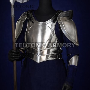 May include: A polished silver metal breastplate, shoulder guards, and arm guards are displayed on a black mannequin. The armor is worn over a dark blue long-sleeved garment. A spear is held in the left hand. The text "TEUTONIC ARMORY" is visible.