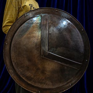 May include: A large, circular, dark metallic shield featuring a prominent V-shaped embossed design across the front surface. The shield has a weathered, textured finish with numerous small indentations and is secured with rivets along the outer rim.