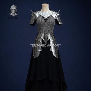 May include: A silver-coloured armour set with shoulder pauldrons, chest and waist plates, and chainmail. The armour is displayed on a mannequin with a black dress underneath. The text "TEUTONIC ARMORY" is visible.