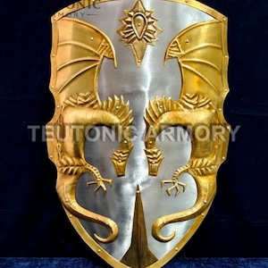 Może przedstawiać: Tarcza w stylu średniowiecznym ze srebrnym środkiem i złotymi wzorami smoków i skrzydeł. Tarcza ma spiczaste dno i ozdobny emblemat na górze. Widoczny jest napis "TEUTONIC ARMORY".