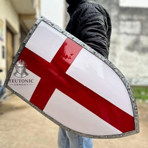 Puede incluir: Un escudo blanco con un diseño de cruz roja, enmarcado con un borde de metal plateado. El escudo tiene el texto "TEUTONIC ARMORY" y un logotipo de casco de caballero. El escudo es sostenido por una persona.