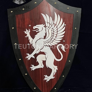 Pode incluir: Um escudo de madeira com centro vermelho e borda preta, adornado com um emblema de grifo branco. O escudo é adornado com tachas douradas e apresenta o texto "TEUTONIC ARMORY".