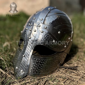 Puede incluir: Un casco medieval plateado con detalles remachados y una prominente guarda nasal. El casco es de metal y tiene una superficie texturizada. Las palabras "TEUTONIC ARMORY" están impresas en el lateral.