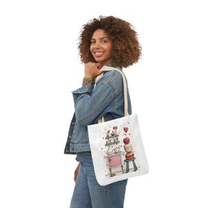 May include: A white tote bag with a whimsical illustration of a pink cabinet, stacked macarons, and a small stool. The bag features a cream-colored strap and is being carried by a person wearing a denim jacket and blue jeans.