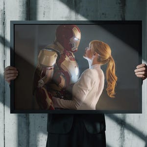 May include: A framed print featuring Iron Man and a woman in a light-colored outfit. The Iron Man suit is red and gold. The woman has long, light-colored hair. The frame is black, and the background is a concrete wall.