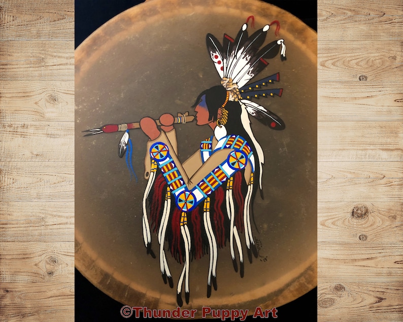 May include: A hand-painted Native American drum featuring a depiction of a person playing a flute. The person is wearing a headdress with feathers and traditional clothing. The drum is brown with a painted design in red, blue, yellow, and white.