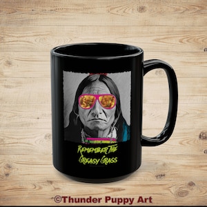 May include: Black ceramic mug featuring a portrait of an Indigenous person wearing pink sunglasses. The image includes the text "Remember the Greasy Grass" in neon green. The mug has a large handle and a glossy finish.
