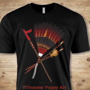May include: Black t-shirt featuring a graphic design of crossed Native American ceremonial items. The design includes a fan, spears, and a gourd, with red, yellow, and brown accents. The text "Thunder Puppy Art" is at the bottom.