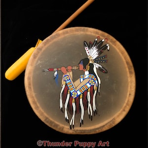 May include: A hand-painted Native American drum featuring a depiction of a person playing a flute. The drum is brown with a white, blue, red, and yellow design. The person is wearing a headdress with feathers. The text "Thunder Puppy Art" is at the bottom of the drum.