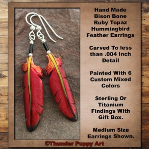 May include: Handmade bison bone hummingbird feather earrings featuring red and yellow paint, ruby topaz accents, and sterling or titanium findings. Text reads: Hand Made Bison Bone Ruby Topaz Hummingbird Feather Earrings, Carved To less than .004 Inch Detail, Painted With 6 Custom Mixed Colors, Sterling Or Titanium Findings With Gift Box. Medium Size Earrings Shown. Thunder Puppy Art.