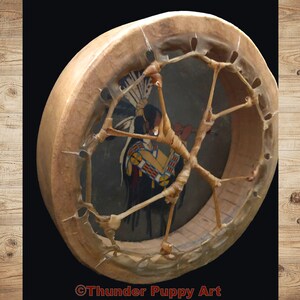 May include: A Native American-style hand drum with a brown leather frame and a painted design of a Native American figure in a headdress. The drum has a woven leather lacing pattern.