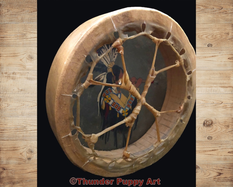 May include: A Native American-style hand drum with a brown leather frame and a painted design of a Native American figure in a headdress. The drum has a woven leather lacing pattern.