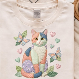 Peut inclure: T-shirt crème avec un motif de chat patchwork brodé. Le chat a des patchs orange, bleus et violets. Des papillons et des cœurs entourent le chat. Le t-shirt a un col rond et des manches courtes.