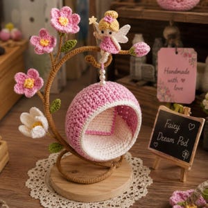 Op de afbeelding: Een handgemaakte gehaakte feeëndroompod in roze en wit, versierd met roze bloemen en een feeënfiguur. De pod hangt aan een bruine gehaakte tak op een houten voet. Een klein schoolbordje leest "Fairy Dream Pod."