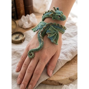 May include: A green and blue crocheted dragon bracelet that wraps around the wrist and features a long tail that extends down to the finger, complete with detailed wings, a textured spine, and small dark eyes on the dragon head.