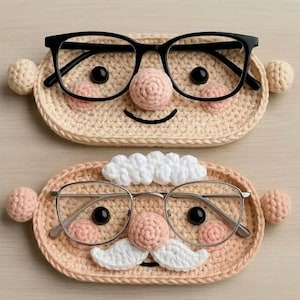 Könnte beinhalten: Zwei handgefertigte Brillenhalter aus Häkelarbeit. Der obere Halter hat eine schwarze Brille, ein beiges Gesicht, rosa Wangen, eine gehäkelte Nase und zwei beige Pompons. Der untere Halter hat eine silberne Brille, ein pfirsichfarbenes Gesicht, einen weißen Schnurrbart und zwei rosa Pompons.