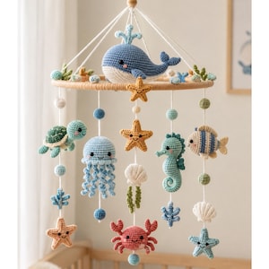 May include: A hanging mobile featuring crocheted sea creatures including a blue whale, green turtle, light blue jellyfish, seahorse, striped fish, crab, and starfish. The figures are suspended from a circular wooden frame with white strings.