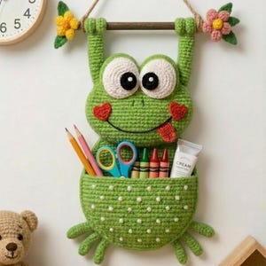 May include: A green crocheted frog-shaped wall organizer with a pocket for storage. The frog has large white eyes, red heart-shaped cheeks, and a smiling mouth. It holds a wooden bar with a rope for hanging. The pocket contains pencils, crayons, and scissors.