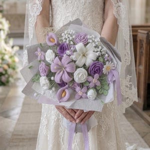 Peut inclure: Un bouquet de fleurs crochetées à la main, composé de roses, de lys, de callas et de marguerites blanches, lavande et violettes. Le bouquet est enveloppé dans du papier gris et noué avec un ruban lavande. Une robe en dentelle blanche est portée.