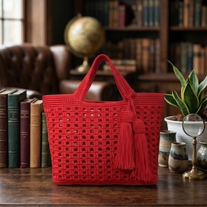 Könnte beinhalten: Eine leuchtend rote Häkeltasche mit rechteckiger Form und Lochmuster. Die Tasche hat zwei Griffe und dekorative Quasten. Die Tasche wird auf einer Holzoberfläche präsentiert, was auf einen handgefertigten Artikel hindeutet.